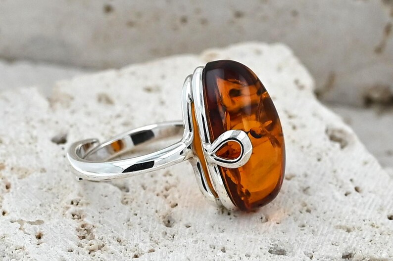 May include: Silver ring with a large, oval amber gemstone. The amber is a warm, translucent orange-brown hue. The ring has a simple, elegant design with a polished silver band and decorative detailing around the stone.