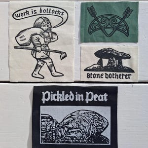May include: A set of four rectangular fabric patches featuring black ink screen-printed designs. One patch shows a person carrying a sack with the text 'work is bollocks'. Another is green with an arrow design. A third shows a stone monument with 'stone botherer'. The last is black with 'Pickled in Peat' and a textured graphic.