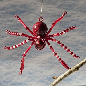 May include: A red and white beaded spider ornament with a shiny, round body and eight legs. The spider is suspended by a silver wire, with a branch in the foreground and a cloudy sky in the background.