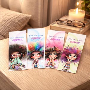 May include: Four colorful bookmarks with illustrations of cartoon girls with large hair. Each bookmark has the text "Just one more CHAPTER" at the top. The bookmarks are on a wooden surface.