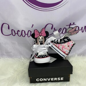 May include: A pair of white Converse trainers with pink and black Minnie Mouse and a pink and white bunny rabbit design. The trainers are decorated with rhinestones and glitter. The trainers are on a white surface with a black Converse box.