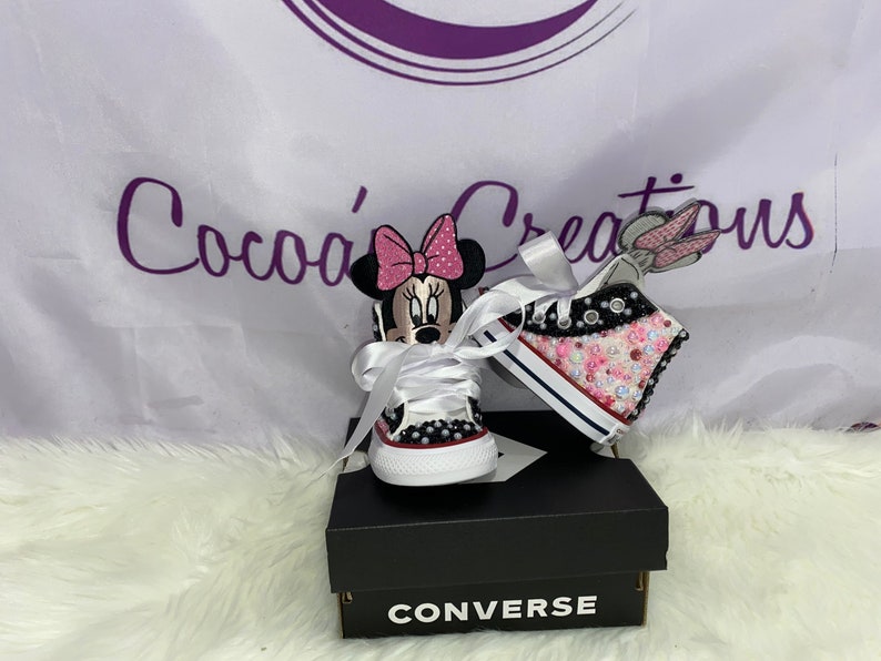 May include: A pair of white Converse trainers with pink and black Minnie Mouse and a pink and white bunny rabbit design. The trainers are decorated with rhinestones and glitter. The trainers are on a white surface with a black Converse box.