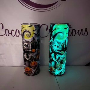 May include: Two stainless steel tumblers with a glow-in-the-dark design featuring a spooky Halloween scene with Jack Skellington from The Nightmare Before Christmas. The tumbler on the left is lit up with a yellow glow, while the tumbler on the right is lit up with a green glow.