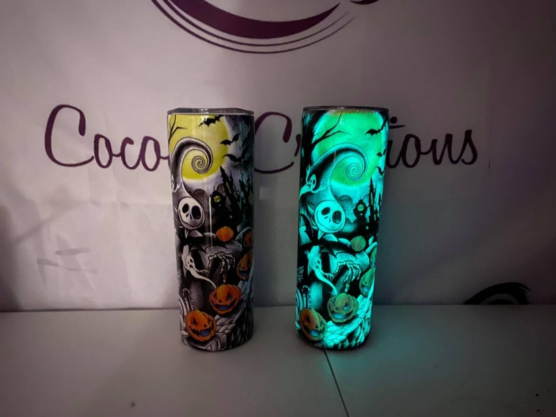 May include: Two stainless steel tumblers with a glow-in-the-dark design featuring a spooky Halloween scene with Jack Skellington from The Nightmare Before Christmas. The tumbler on the left is lit up with a yellow glow, while the tumbler on the right is lit up with a green glow.