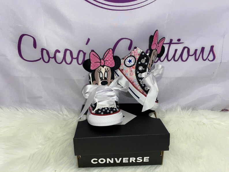 May include: A pair of white Converse trainers with a pink and black Minnie Mouse design. The trainers are decorated with rhinestones and have white satin laces. The trainers are in a box with the Converse logo.