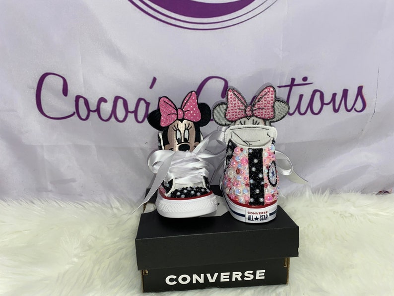 May include: A pair of white Converse trainers with pink and black Minnie Mouse designs. The trainers are decorated with rhinestones and have white laces. The trainers are in a black box with the word "CONVERSE" printed on the front.