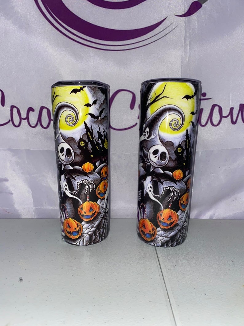 May include: Two black and white stainless steel tumblers with a Halloween themed design featuring Jack Skellington from The Nightmare Before Christmas. The design includes a yellow moon, a spooky tree, and pumpkins.
