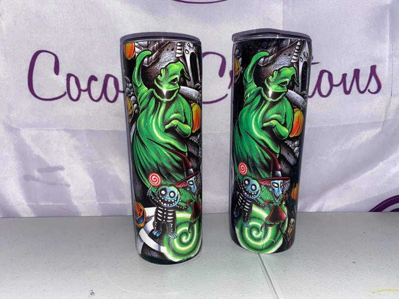 May include: Two black and green stainless steel tumblers with a Halloween theme. The tumblers feature a green ghost with a black hat and a swirl design. The ghost is surrounded by smaller characters with black hats and green faces.