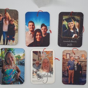 May include: Six white rectangular photo ornaments with red string hangers. Each ornament features a different family photo. The photos include a family of three, a family of four, a graduation photo, a family of two, and a family of three.