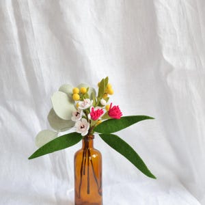 May include: A small, amber glass bottle contains a felt flower arrangement. The arrangement features green leaves, white and pink flowers, and yellow accents. The background is a white, textured fabric.