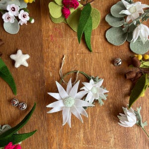 May include: A collection of felt craft supplies including white flower ornaments with pale green centres, green felt leaves, a white felt star, small silver jingle bells, and brown felt buds arranged on a wooden surface for decorative projects.