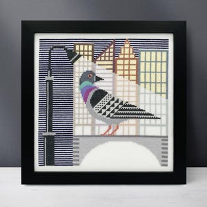 May include: A cross-stitch pattern of a pigeon perched on a bridge in front of a city skyline. The pigeon is gray with purple and white markings. The city skyline is made up of buildings with different colored windows.