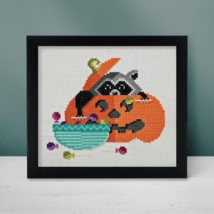 May include: Cross stitch pattern featuring a raccoon peeking out of a pumpkin with a bowl of candy in front of it.
