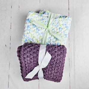 May include: Two folded crochet blankets, one with a speckled pattern of white, blue, and green, and the other in solid purple. Each blanket is tied with a ribbon, the top one with a green and white ribbon, and the bottom one with a white ribbon.