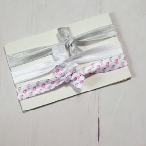 May include: Three hair ties on a white card. One hair tie is silver, one is white, and one is white with a pink and white bird pattern.