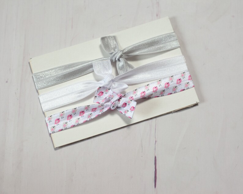 May include: Three hair ties on a white card. One hair tie is silver, one is white, and one is white with a pink and white bird pattern.