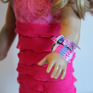 May include: A pink ruffled dress with a colourful fabric wristband. The wristband is tied in a bow.