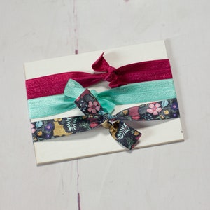 May include: Three hair ties on a white background. One is a dark red, one is a light blue, and one is a dark blue with a floral pattern.