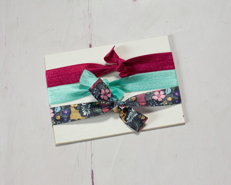 May include: Three hair ties on a white background. One is a dark red, one is a light blue, and one is a dark blue with a floral pattern.