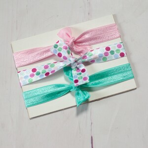 May include: Three hair ties on a white card. One hair tie is pink, one is turquoise, and one is white with pink, green, and turquoise polka dots.