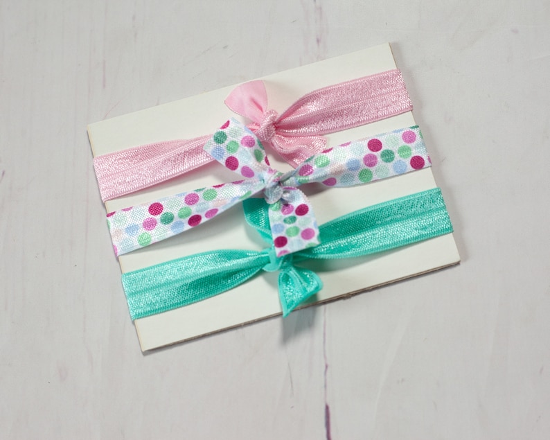 May include: Three hair ties on a white card. One hair tie is pink, one is turquoise, and one is white with pink, green, and turquoise polka dots.