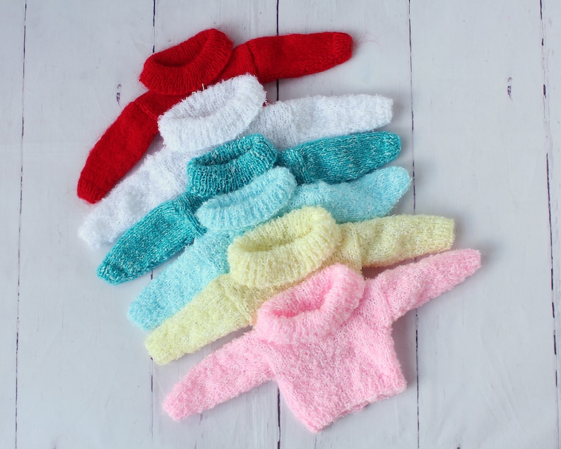May include: A stack of seven colorful knitted sweaters in various shades of red, white, teal, light blue, yellow, and pink. The sweaters are arranged in a rainbow pattern, with the red sweater on top and the pink sweater on the bottom.