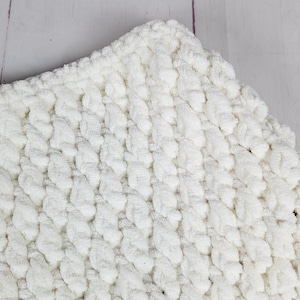 May include: Close-up of a soft, white blanket with a textured, braided pattern. The blanket is made of a plush material, and the corner is visible, showing the thickness and detail of the weave. The blanket is a cozy home decor item.