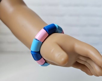 May include: A vibrant bracelet with cylindrical beads in blue and pink hues. The bracelet is displayed on a wrist, highlighting the alternating colours. The beads are smooth and uniform, creating a playful and stylish accessory. The bracelet's circumference is approximately 18 cm.