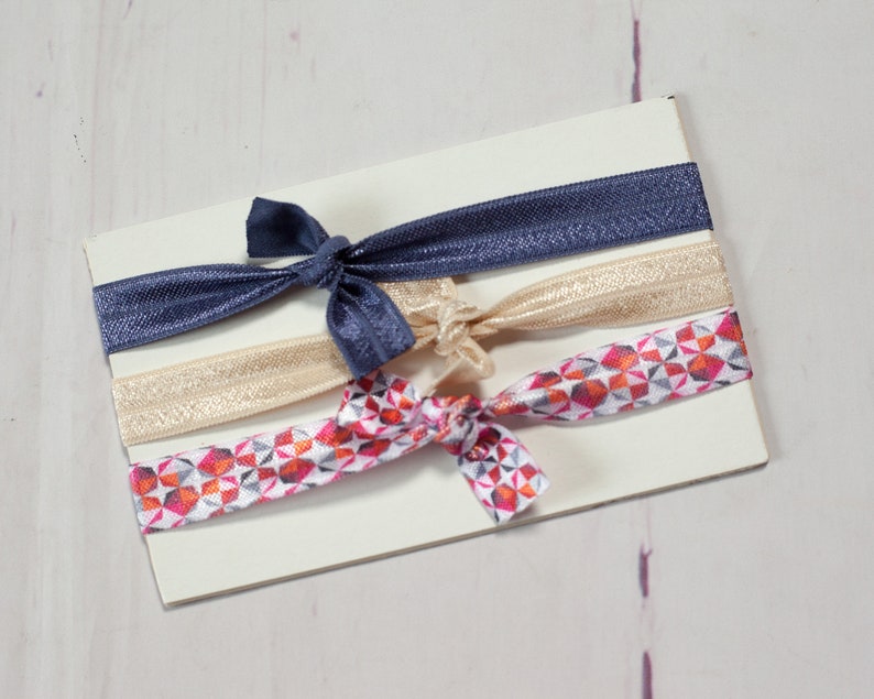 May include: A set of three hair ties, one navy blue, one gold, and one with a geometric pattern in shades of pink, orange, and white.