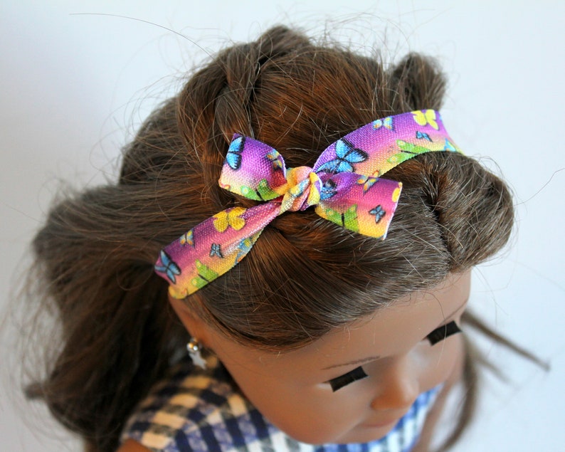 May include: A doll with brown hair wearing a headband with a rainbow and butterfly pattern.
