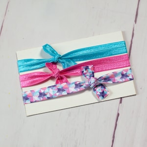 May include: Three hair ties, one with a blue solid colour, one with a pink solid colour, and one with a pink, blue and white geometric pattern.