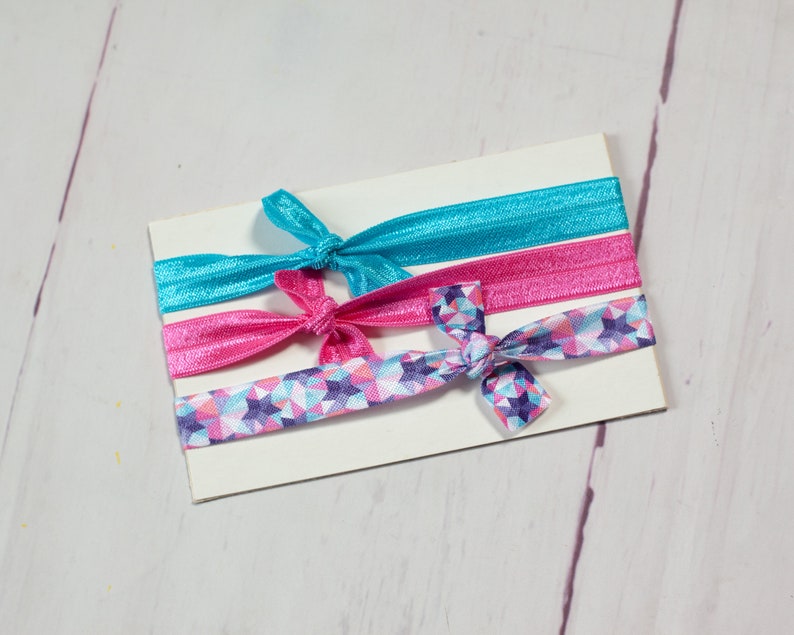 May include: Three hair ties, one with a blue solid colour, one with a pink solid colour, and one with a pink, blue and white geometric pattern.