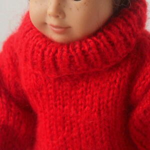 May include: A red knitted turtleneck sweater for a doll. The sweater is made of a soft, fuzzy yarn.