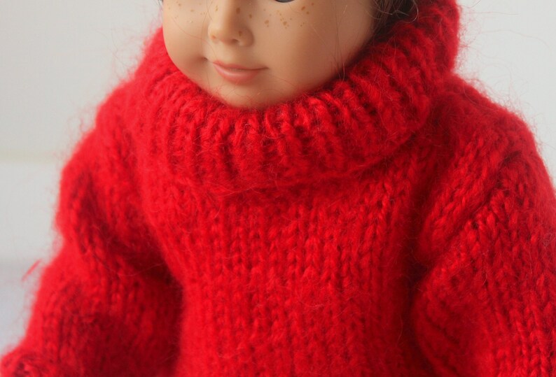 May include: A red knitted turtleneck sweater for a doll. The sweater is made of a soft, fuzzy yarn.