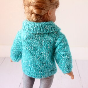 May include: A turquoise knitted sweater with a white fleck pattern for a doll. The sweater has a collared neckline and long sleeves.