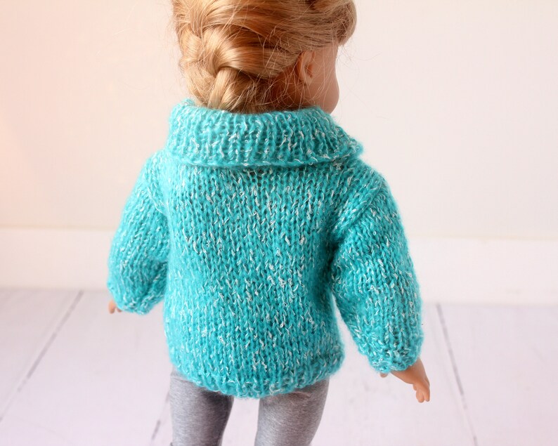 May include: A turquoise knitted sweater with a white fleck pattern for a doll. The sweater has a collared neckline and long sleeves.
