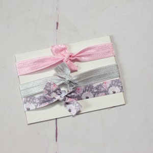 May include: Three hair ties on a white card. One is pink, one is silver, and one is a floral print with pink and grey flowers.