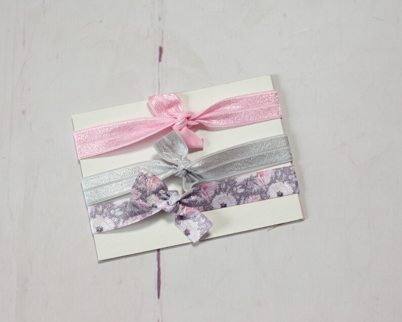 May include: Three hair ties on a white card. One is pink, one is silver, and one is a floral print with pink and grey flowers.