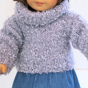 May include: A gray fuzzy turtleneck sweater for a doll. The sweater has a loose fit and a wide, folded turtleneck collar.