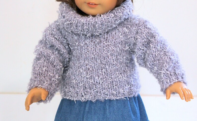 May include: A gray fuzzy turtleneck sweater for a doll. The sweater has a loose fit and a wide, folded turtleneck collar.