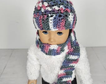 May include: A doll wearing a handmade crochet hat and scarf set. The hat and scarf feature a colorful pattern with shades of blue, pink, white, and gray. The doll is also wearing a white sweater.