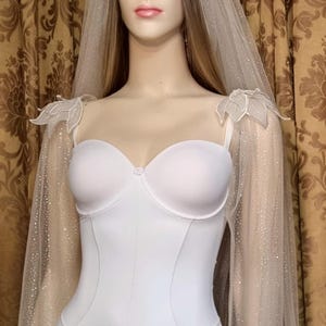 May include: A white bridal bodysuit with a sweetheart neckline and delicate straps. The bodysuit is paired with a sheer, sparkling veil that drapes over the shoulders. The mannequin is displayed in front of a gold patterned curtain.