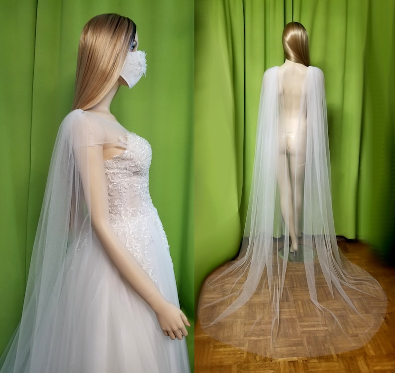 May include: A white tulle bridal cape with a long train. The cape is made of soft, sheer fabric and has a delicate, flowing design.