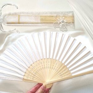 May include: A white folding fan with a wooden handle and intricate designs. The fan is made of white fabric and has a delicate, feminine look.