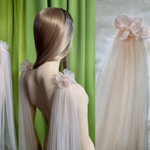 May include: Two sheer tulle bridal capes with delicate pink flower embellishments on the shoulders. The capes are draped over a mannequin and are shown from the back.