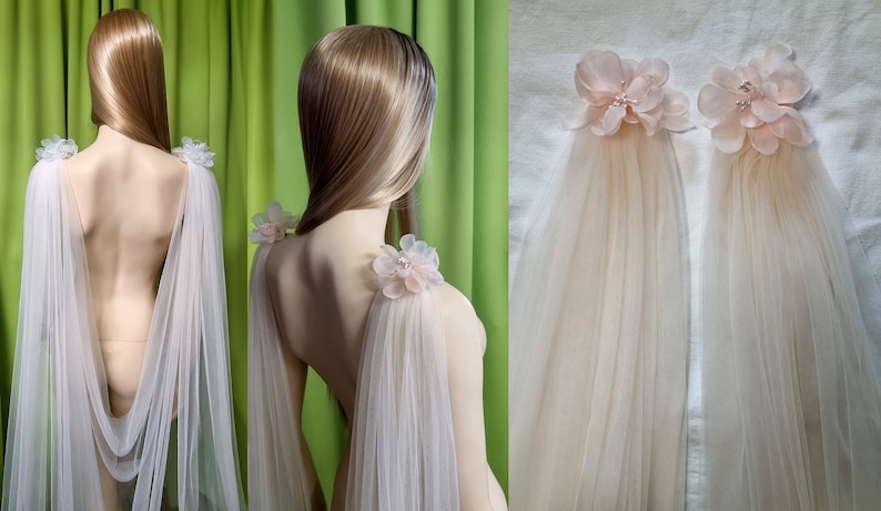 May include: Two sheer tulle bridal capes with delicate pink flower embellishments on the shoulders. The capes are draped over a mannequin and are shown from the back.