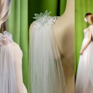 May include: A white tulle bridal cape with delicate floral embellishments on the shoulders. The cape is shown on a mannequin with a green background.