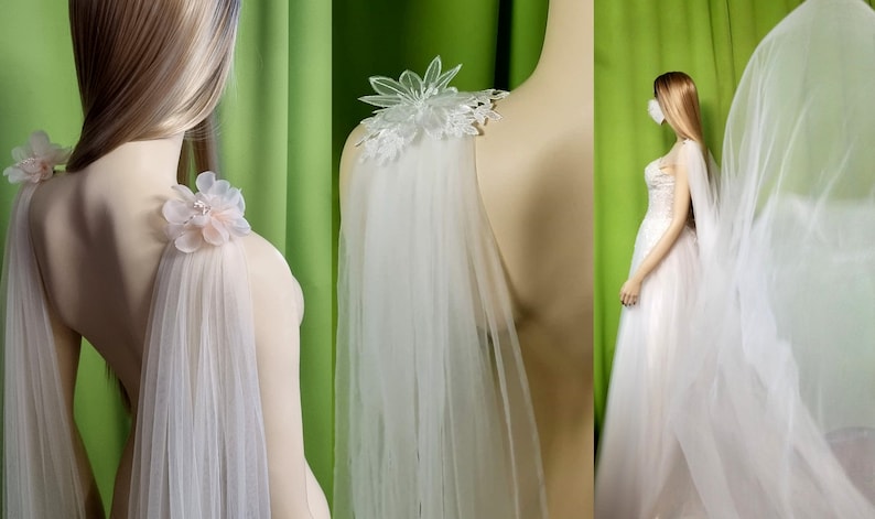 May include: A white tulle bridal cape with delicate floral embellishments on the shoulders. The cape is shown on a mannequin with a green background.