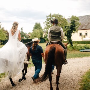 May include: A bride and groom riding horses in a rural setting. The bride is wearing a white wedding dress and the groom is wearing a brown vest and a cowboy hat. A photographer is capturing the moment.