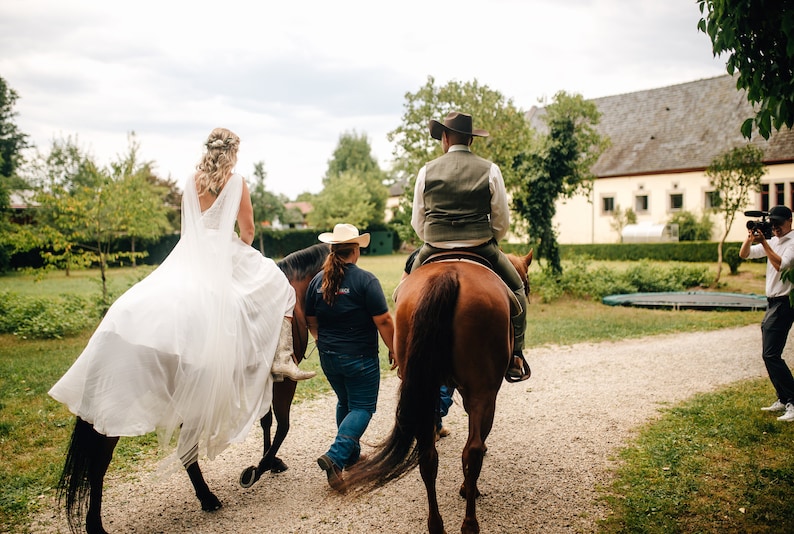 May include: A bride and groom riding horses in a rural setting. The bride is wearing a white wedding dress and the groom is wearing a brown vest and a cowboy hat. A photographer is capturing the moment.